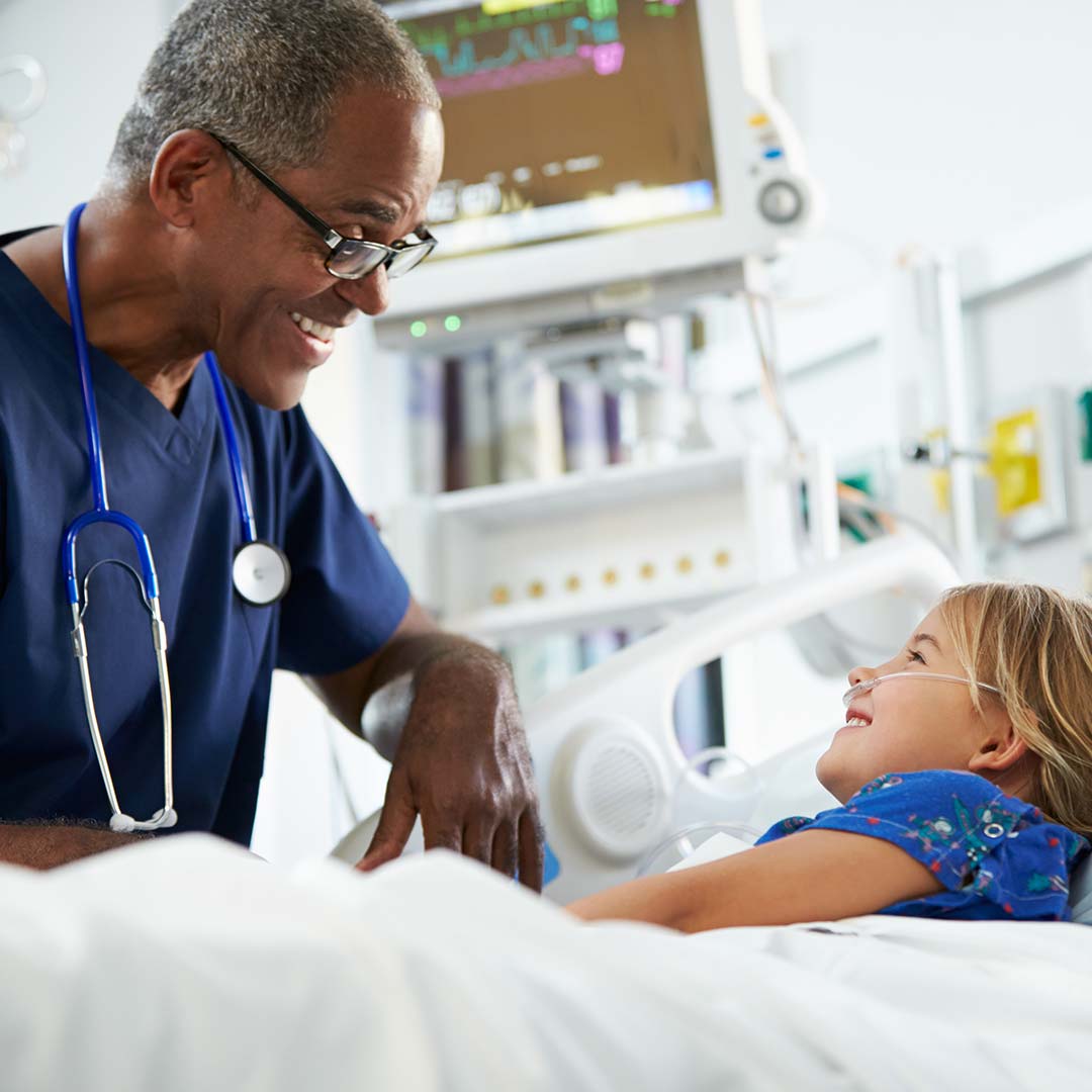 A patient girl is lying on the bed and a doctor is standing nearby. They are smiling at each other and looking happy.